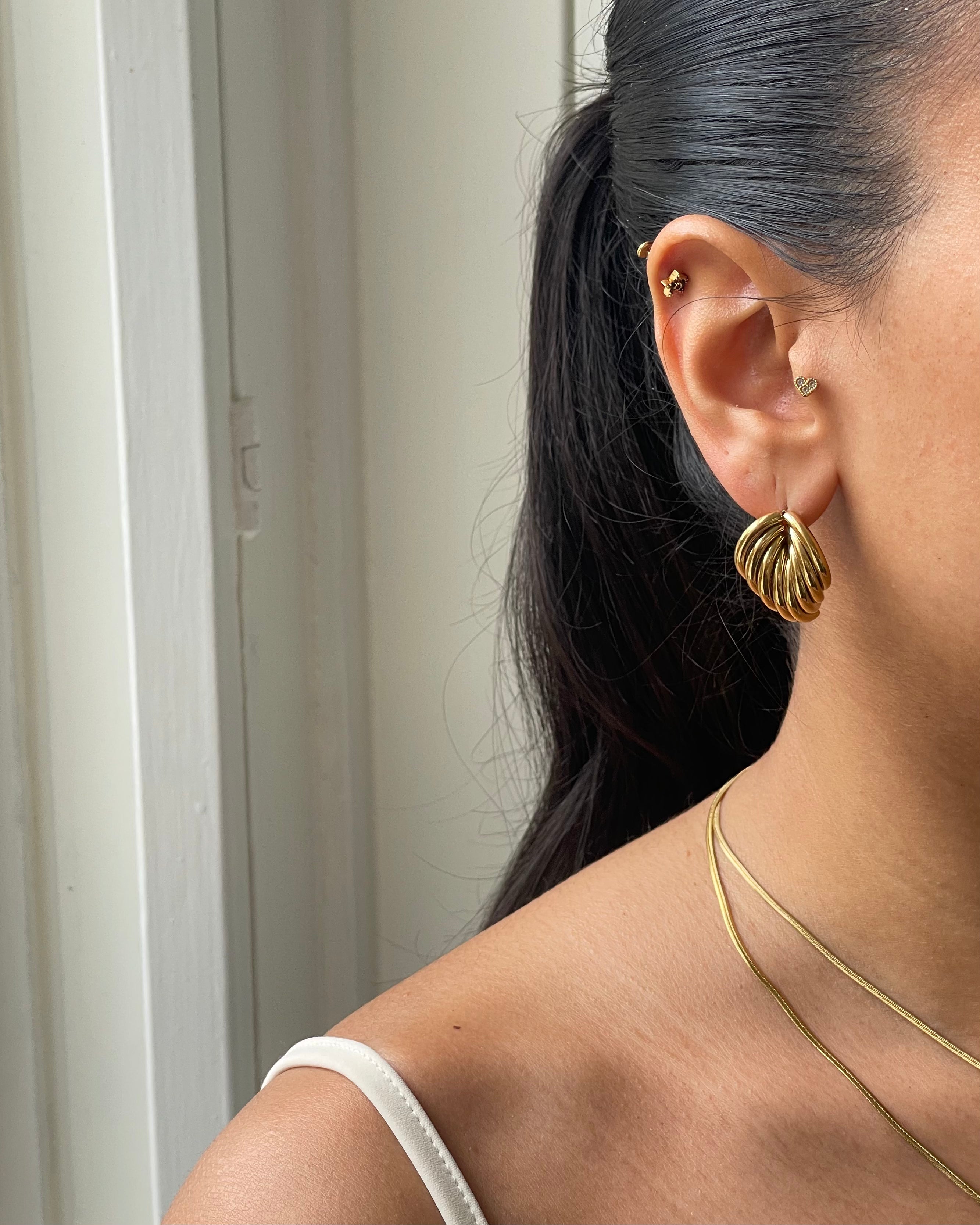Nahaufnahme Ohr mit elegantem goldfarbenem Ohrschmuck, kleinen Herz- und Blüten-Piercings, dunklem Haar